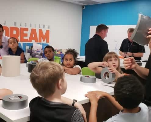Group of 1st and 2nd grade youth with 3M volunteers watches as man demonstrates how to make wallets out of duct tape.
