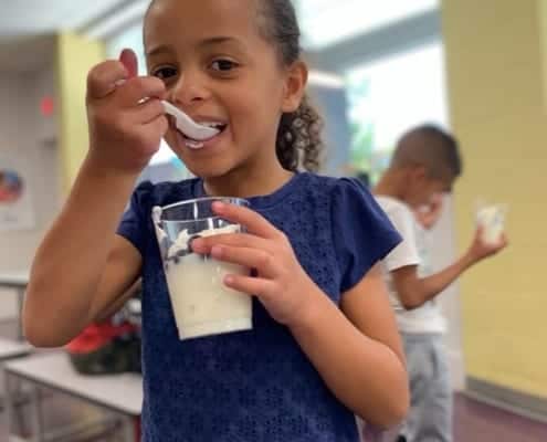 Girl smiles at camera in cafeteria, while eating healthy yogurt parfait snack made with local ingredients for Oregon's summer meals week challenge.
